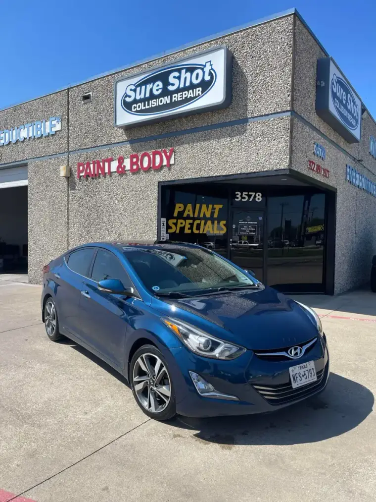 A repaired blue Hyundai Elantra parked in front of the Sure Shot Collision Repair building sign